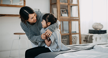 Mother and young daughter sitting on edge of bed, hugging